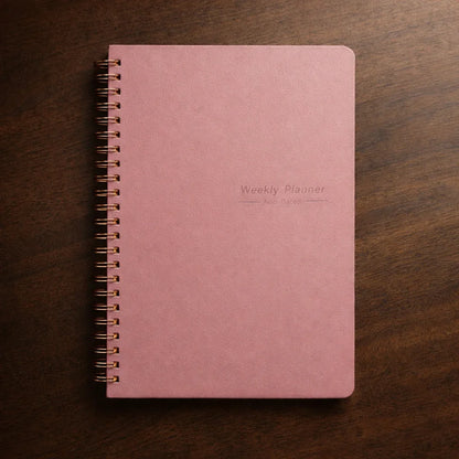 Pink weekly planner with gold spiral binding, non-dated, on wood surface